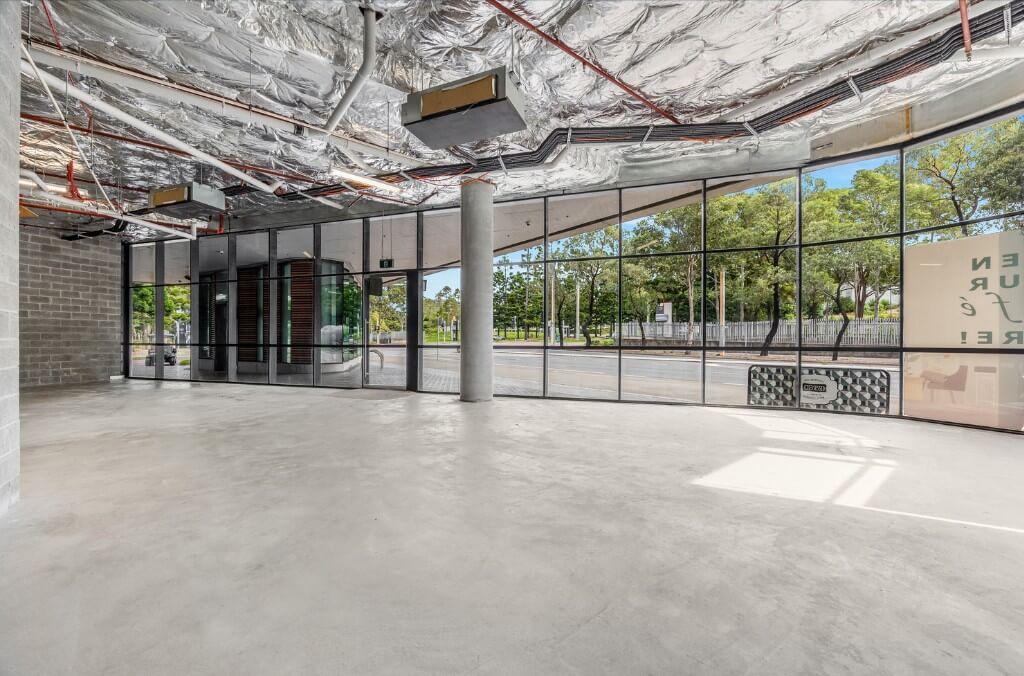 An empty commercial space with large floor-to-ceiling windows, exposed ceiling ducts, and concrete flooring, overlooking a tree-lined street.