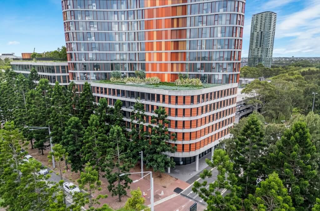 Another angle of outside the building. A modern multi-story building with distinctive orange and glass facade surrounded by green trees, and curved architectural elements.